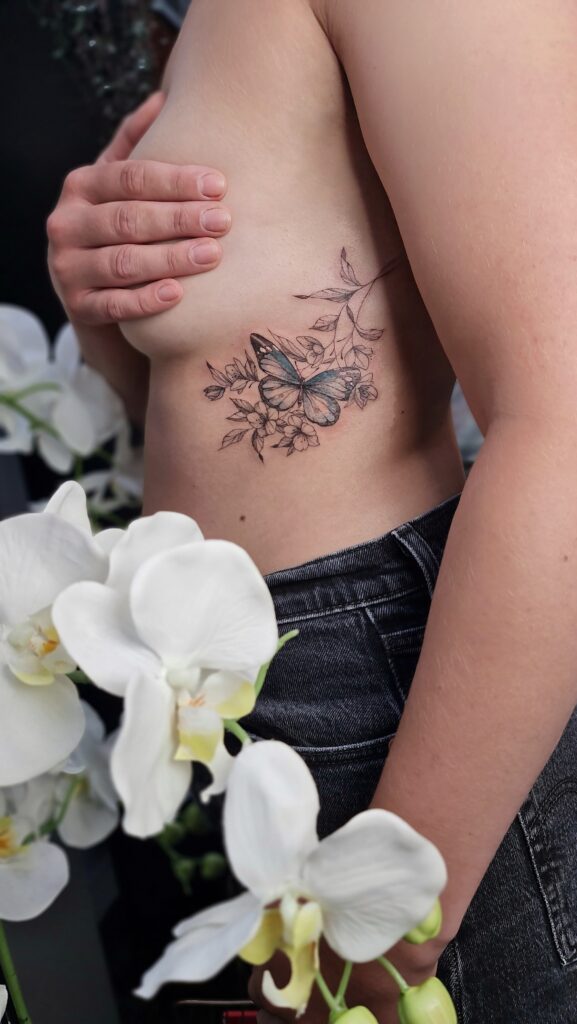 A soft blue butterfly rests among delicate floral branches in this elegant rib tattoo.
