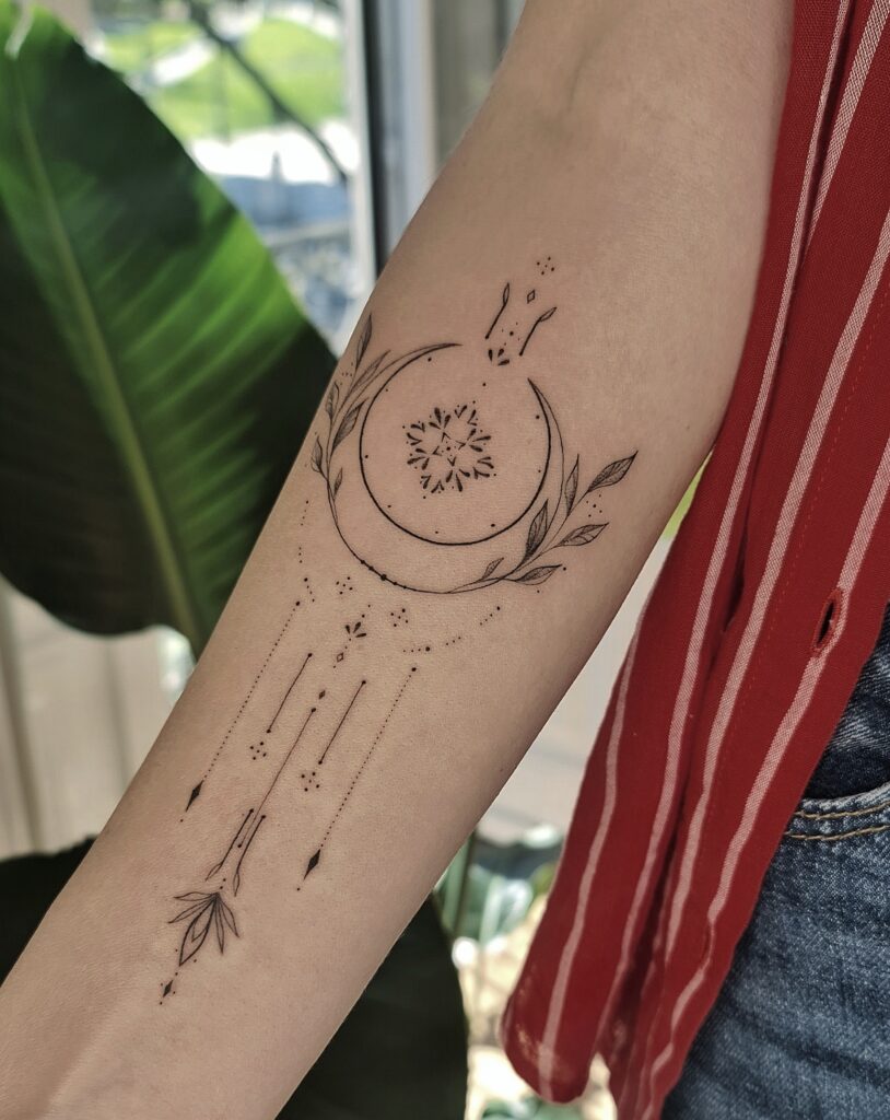 A crescent moon cradling a mandala center, wrapped in botanicals and trailing delicate arrows and dotwork down the forearm.