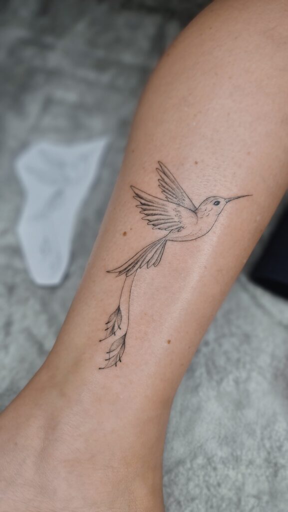 A delicate hummingbird hovers mid-flight, its tail feathers gracefully transforming into trailing leaves in this elegant ankle piece.