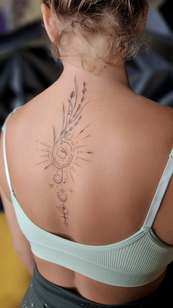 A fine-line spine tattoo featuring a treble clef radiating sunburst lines, intertwined with botanical sprigs, moon phases, music notes, and mystical symbols.