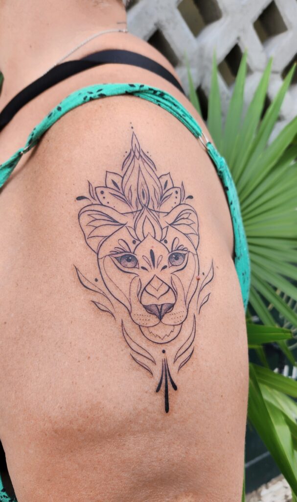 A fine-line shoulder tattoo of a lioness face crowned with mandala-inspired floral petals, framed by flowing botanical accents and dotwork details.