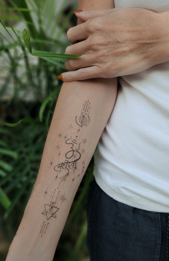 A fine-line forearm tattoo combining Devanagari script reading "Om Mahadevi," a Cho Ku Rei Reiki symbol, scattered star sparkles, and a geometric star tetrahedron (merkaba) connected by dotted lines.