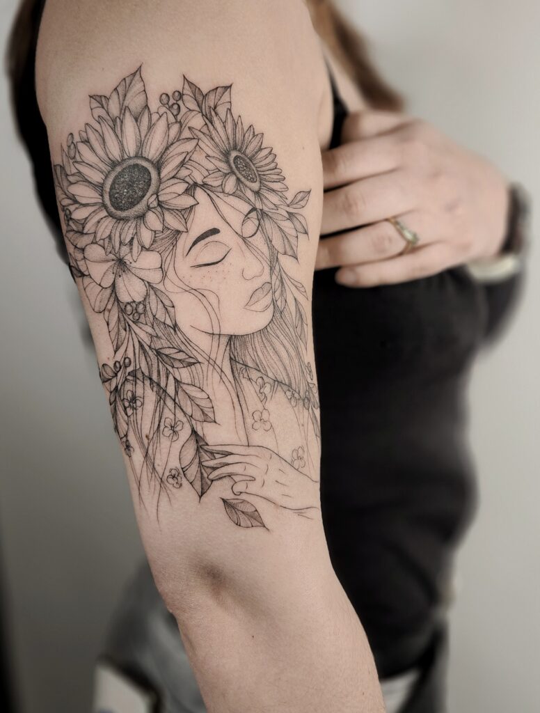 A detailed fine-line upper arm tattoo of a serene woman with closed eyes, her hair and crown overflowing with sunflowers, leaves, and small wildflowers as she gently touches her chest.