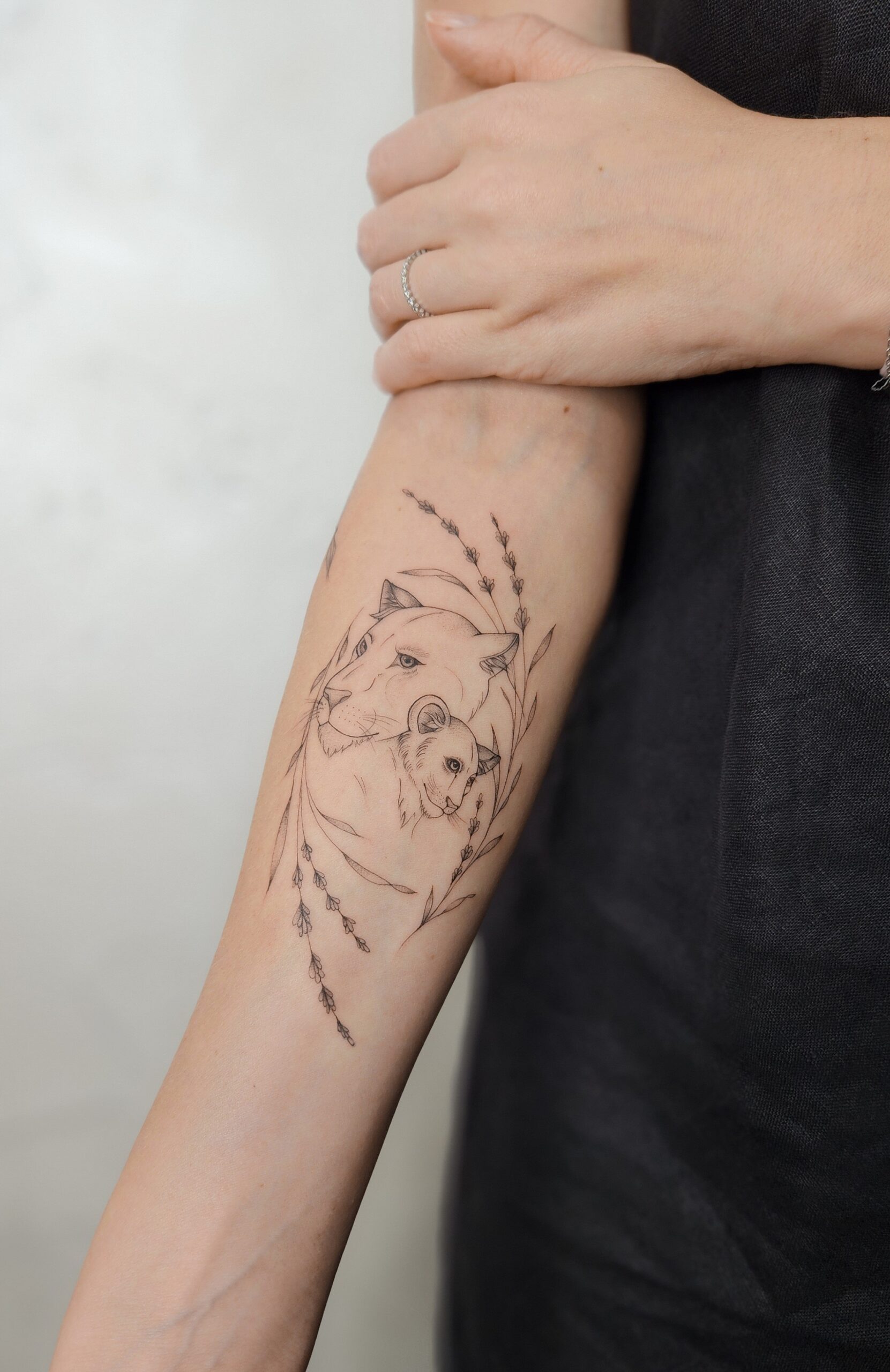 A tender fine-line forearm tattoo of a lioness nuzzling her cub, framed by delicate sprigs of lavender and wispy leaves.