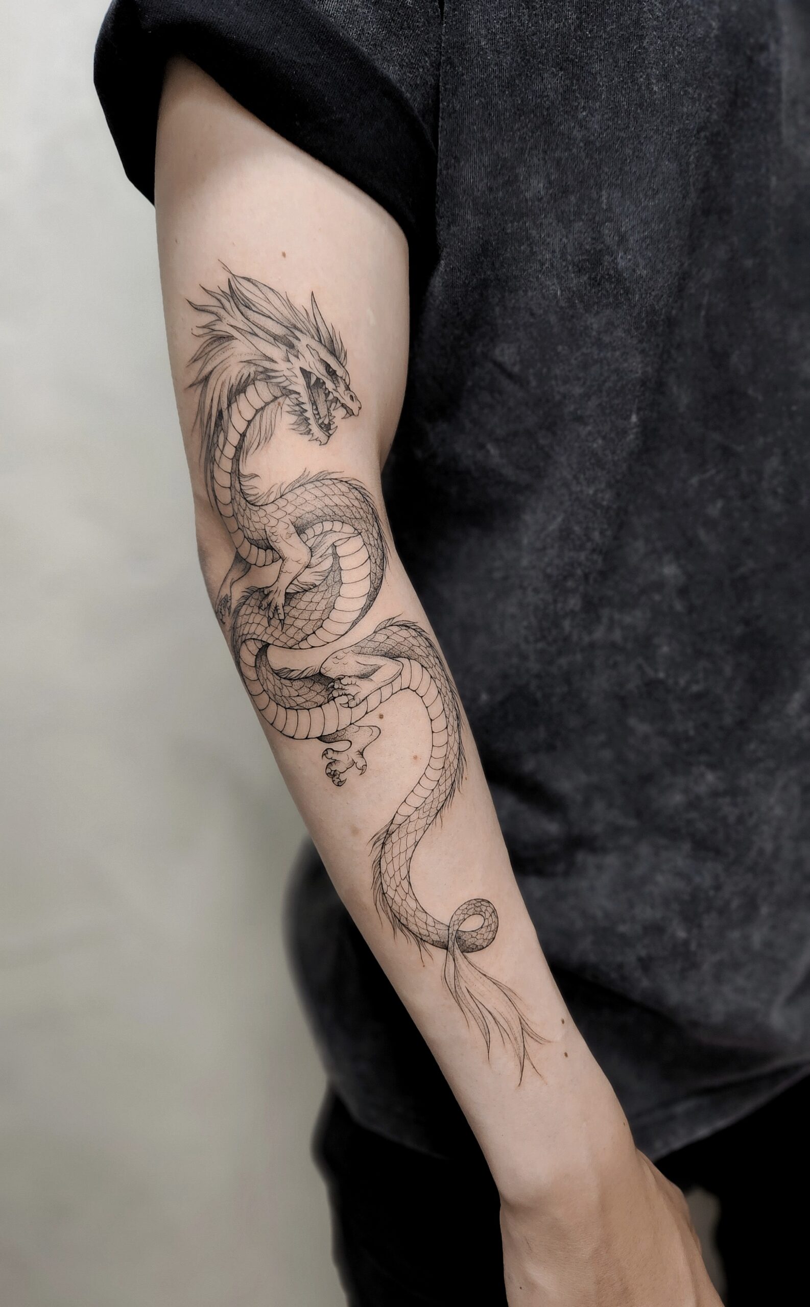 A detailed fine-line tattoo of a sinuous Eastern dragon coiling down the entire arm, from bicep to wrist, with intricately rendered scales, clawed feet, flowing whiskers, and a wispy tail fin.