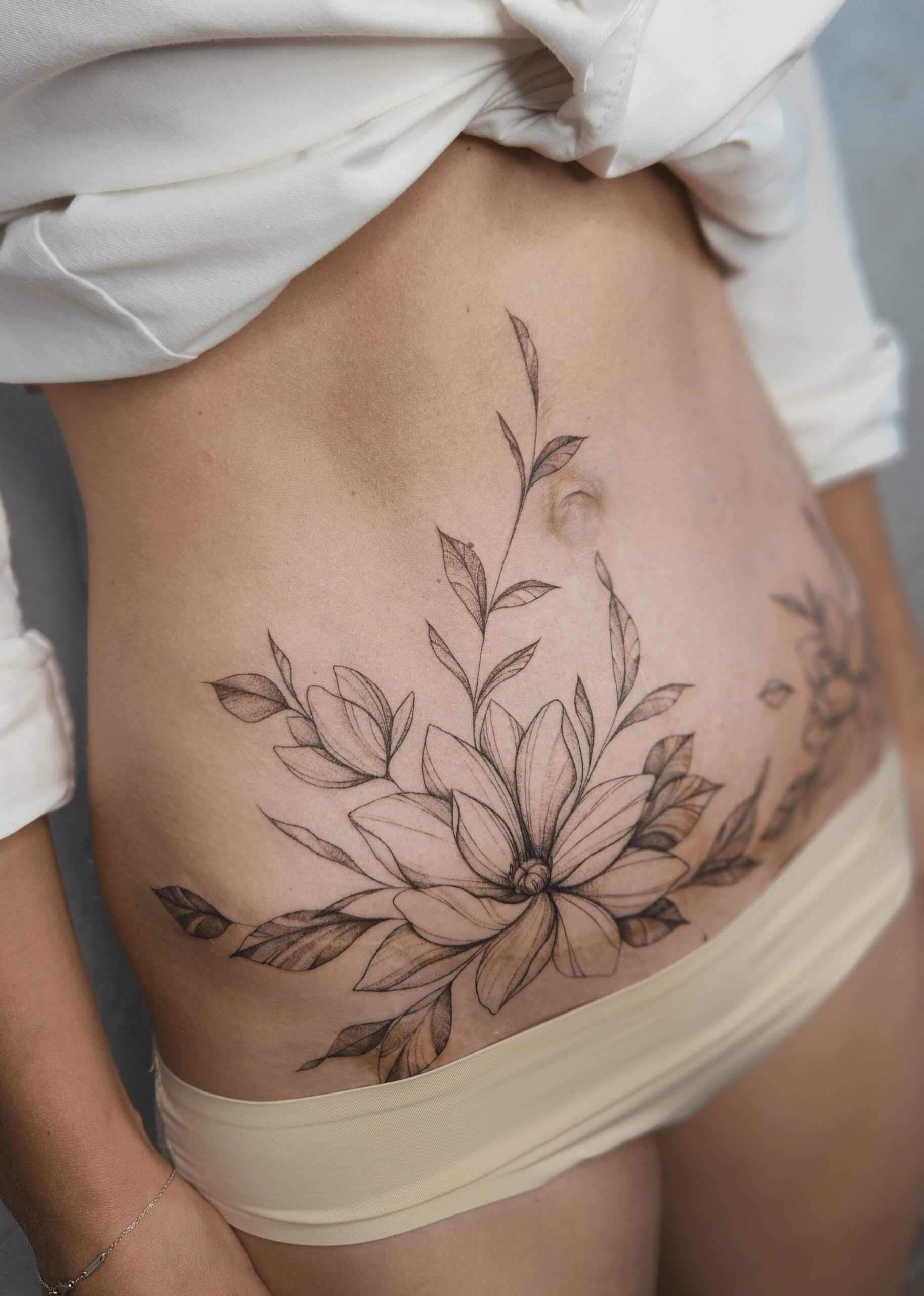 A large fine-line stomach tattoo featuring a lush, open dahlia bloom surrounded by cascading leaves and a slender branch climbing toward the navel.