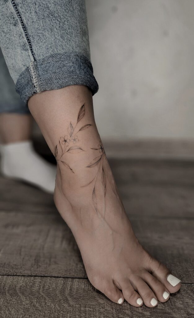 A wispy fine-line tattoo of trailing leaves and small blossoms wrapping organically around the ankle and down onto the top of the foot.