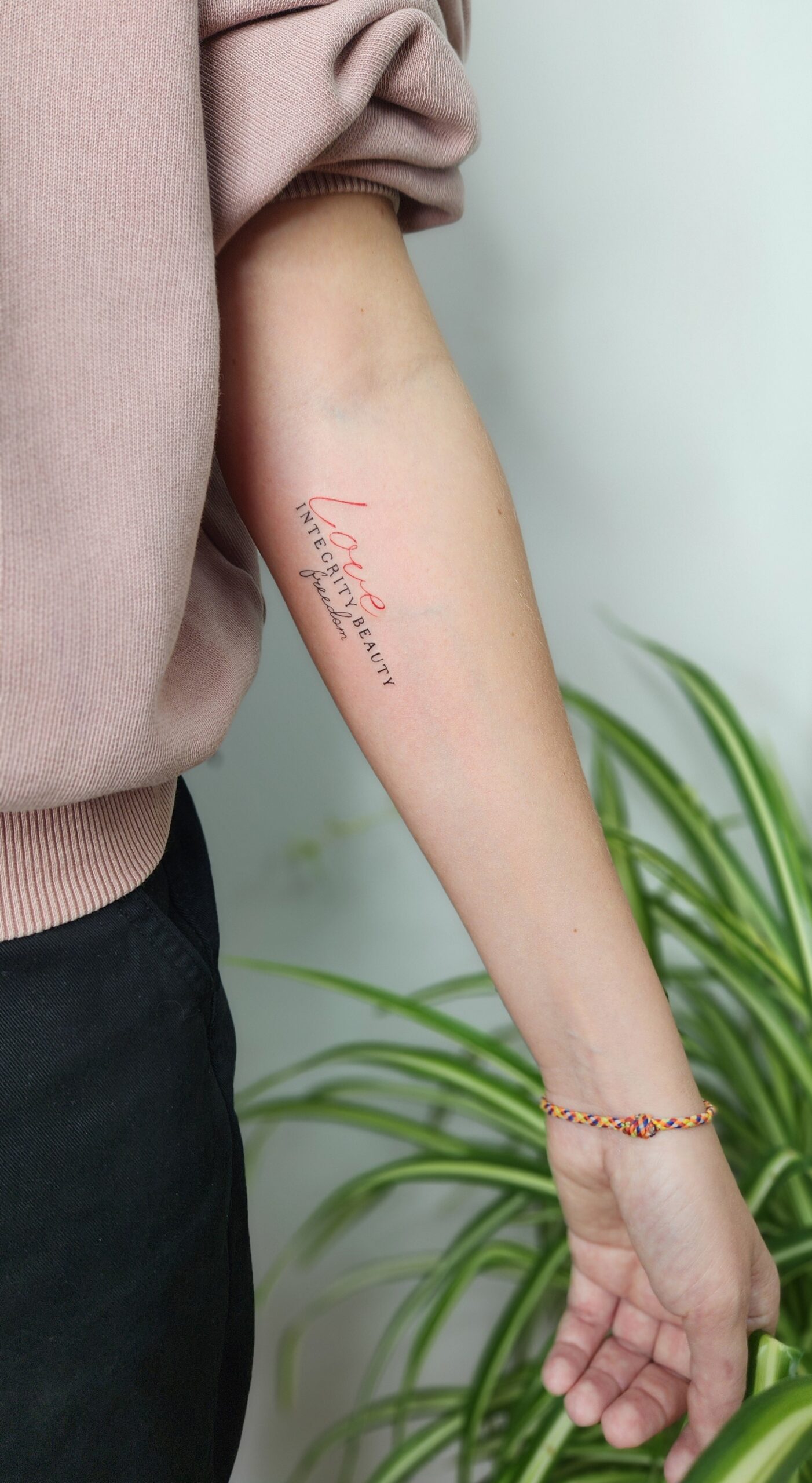A delicate forearm tattoo featuring the words “love” in red ink and “integrity,” “beauty,” and “freedom” in black, arranged in a cascading handwritten script.