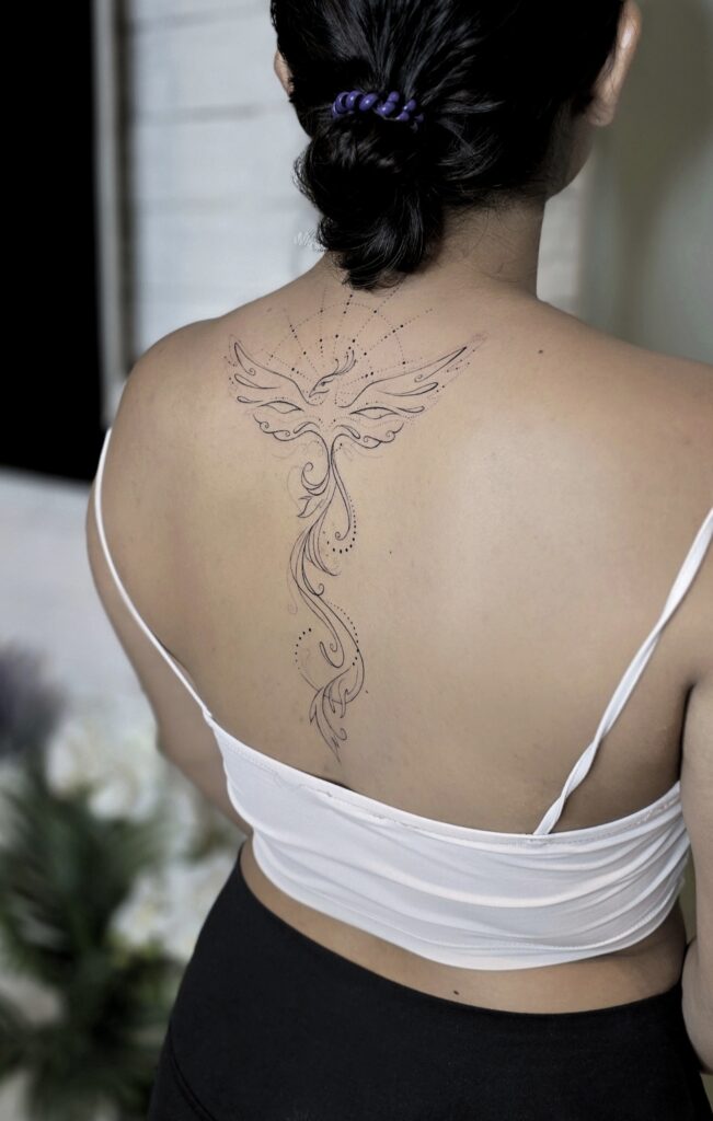 A graceful fine-line phoenix tattoo rising along the spine, with outstretched wings framed by geometric dotwork and a flowing ornamental tail cascading down the back.