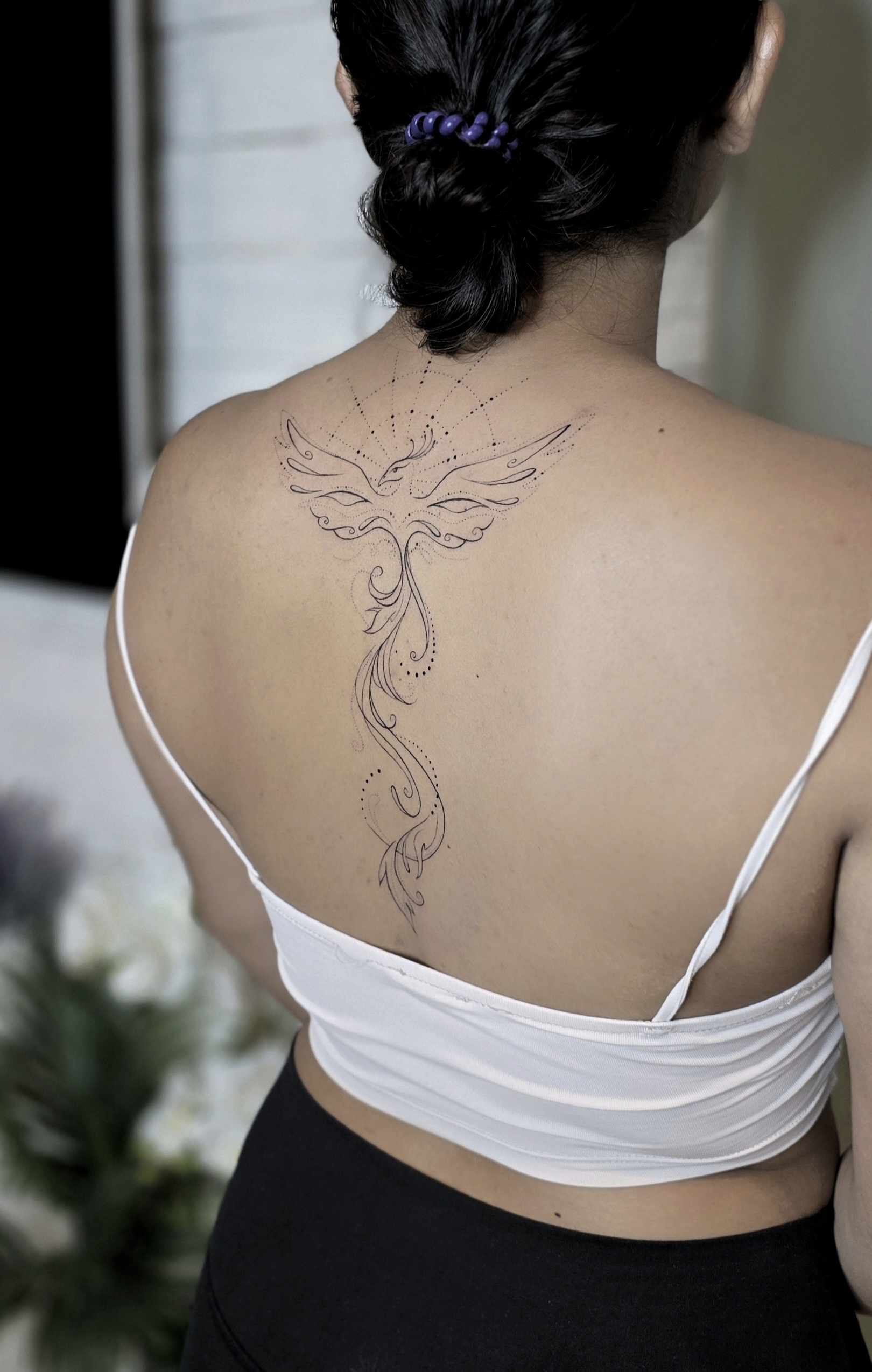 A graceful fine-line phoenix tattoo rising along the spine, with outstretched wings framed by geometric dotwork and a flowing ornamental tail cascading down the back.
