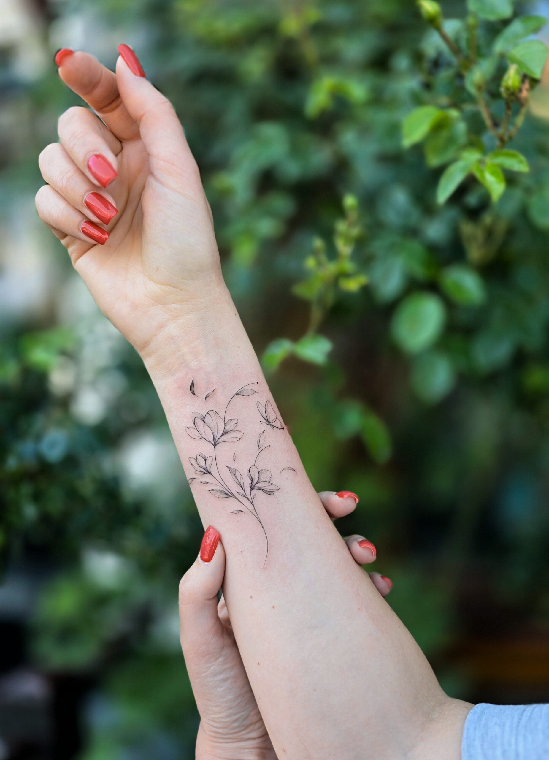 A fine-line wrist tattoo of a slender magnolia branch with open blooms, buds, and a few drifting petals, rendered in soft black ink.