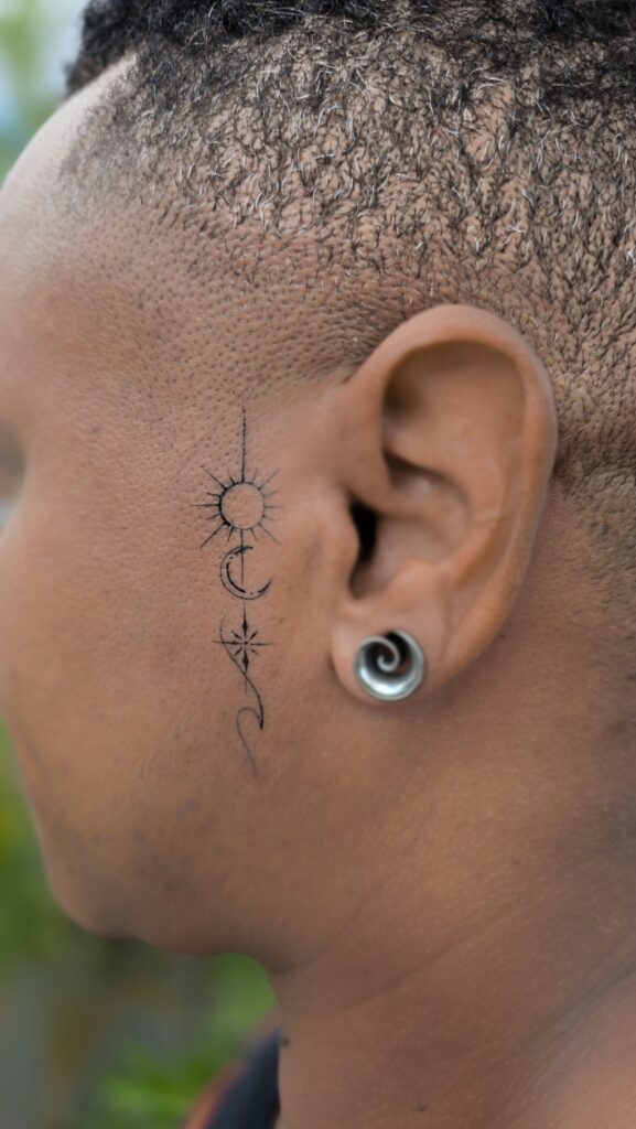 A tiny vertical tattoo behind the ear featuring a radiating sun, a crescent moon with dots, and a compass-style star connected by a fine ornamental line.