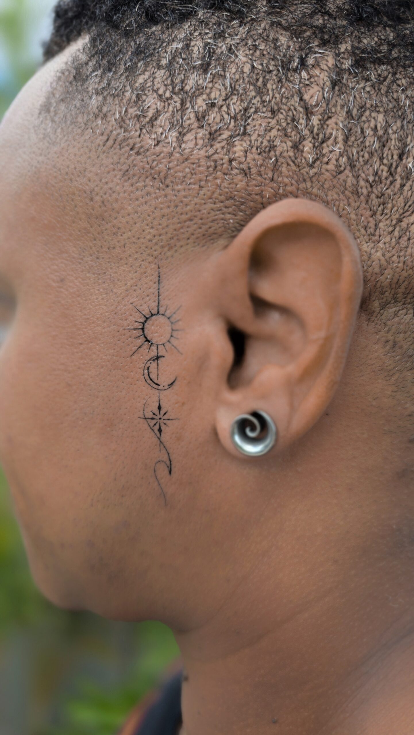 A tiny vertical tattoo behind the ear featuring a radiating sun, a crescent moon with dots, and a compass-style star connected by a fine ornamental line.